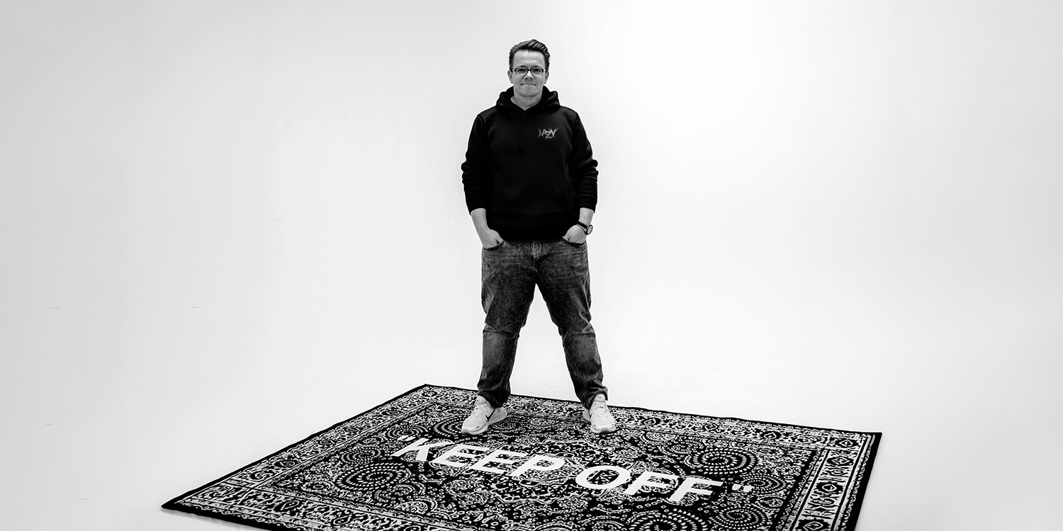 Sebastian Seelig steht in Kapuzenpulli und Jeans auf einem Teppich mit der Aufschrift KEEP OFF